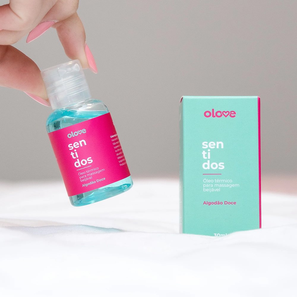 M�o de pessoa segurando o frasco do �leo para Massagem Sensual Olove Sentidos Algod�o Doce 30ml, com a caixa ao fundo