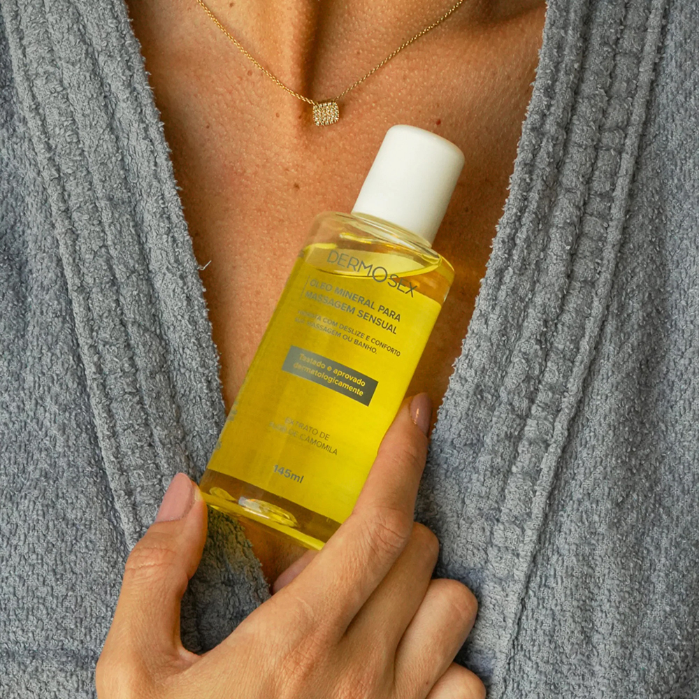 Frasco do �leo Mineral para Massagem Sensual DERMOSex 145ml, sendo segurado por uma m�o, com robe de banho ao fundo.