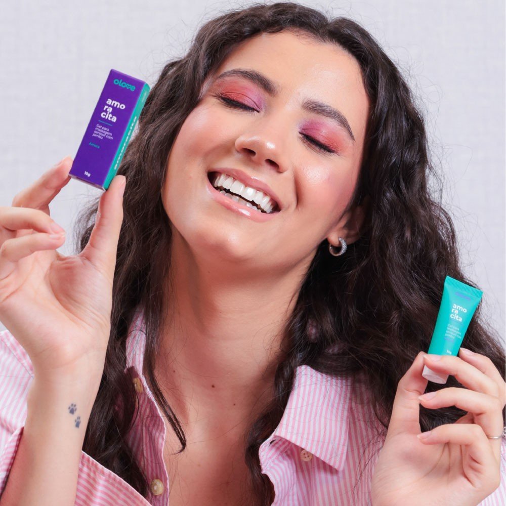 Modelo feminina sorrindo e segurando o Gel Vibrador L�quido Beij�vel Amoracita 10ml e sua embalagem, demonstrando o tamanho pr�tico do produto.