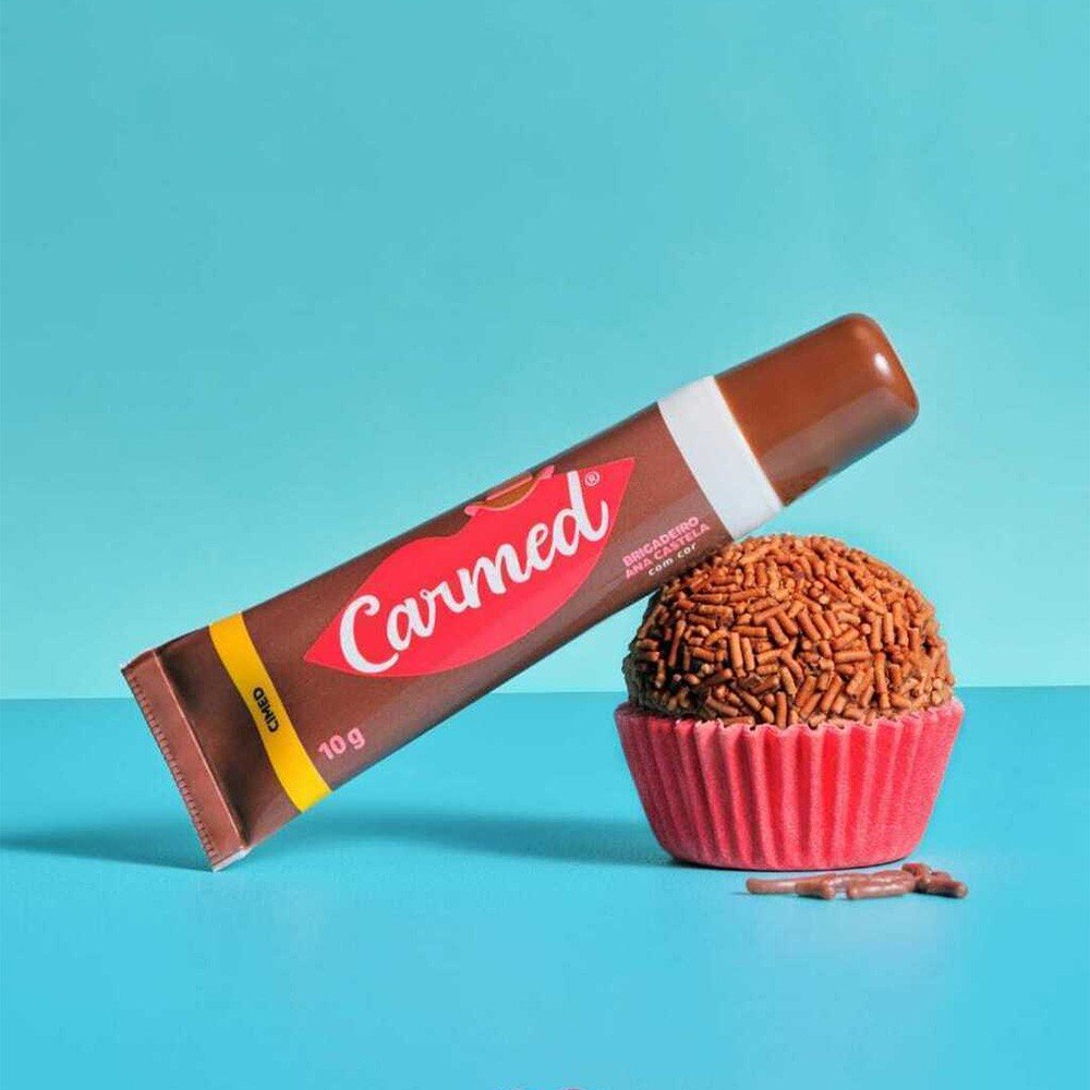 Hidratante Labial Carmed Brigadeiro bisnaga ao lado de um doce de brigadeiro em fundo azul