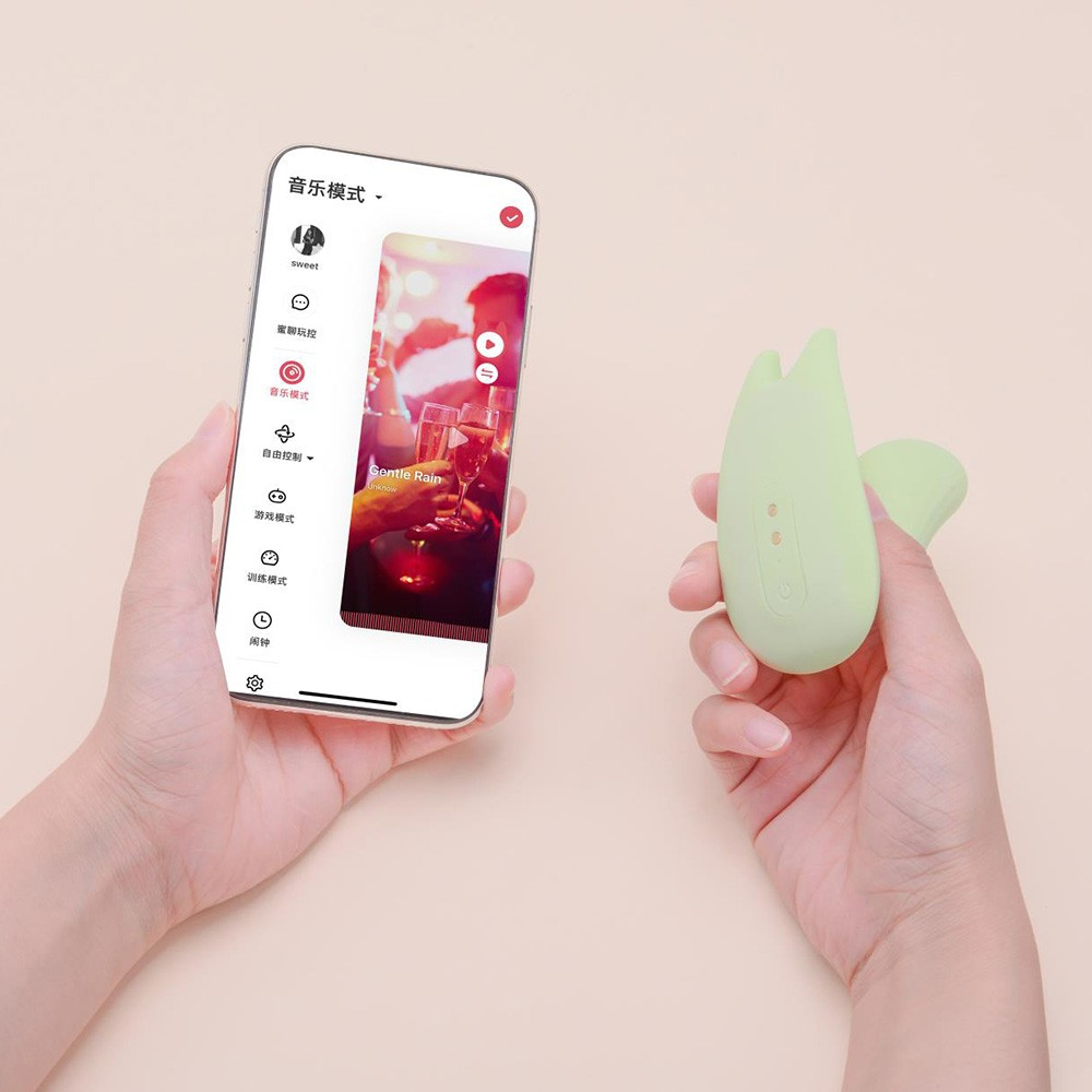 Vibrador Duplo em Silicone Recarreg�vel Verde, sendo segurado por m�os femininas ao lado de um smartphone com o aplicativo
